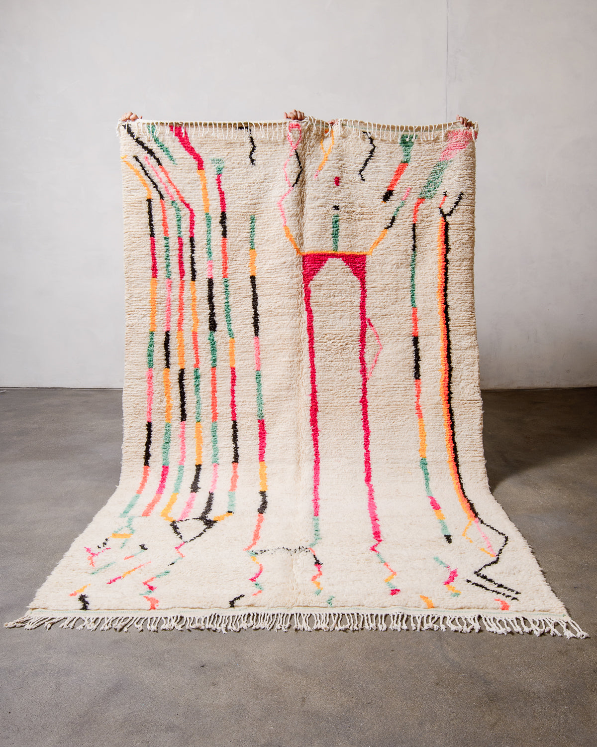Berberlin Azilal rug made of fluffy cream wool and colourful design, held up in front of a white wall and cement floor.